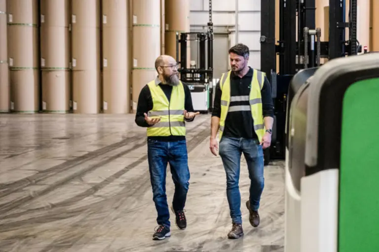 Deux personnes échangeant à proximité d'un chariot de manutention dans un environnement industriel avec des bobines stockées verticalement.